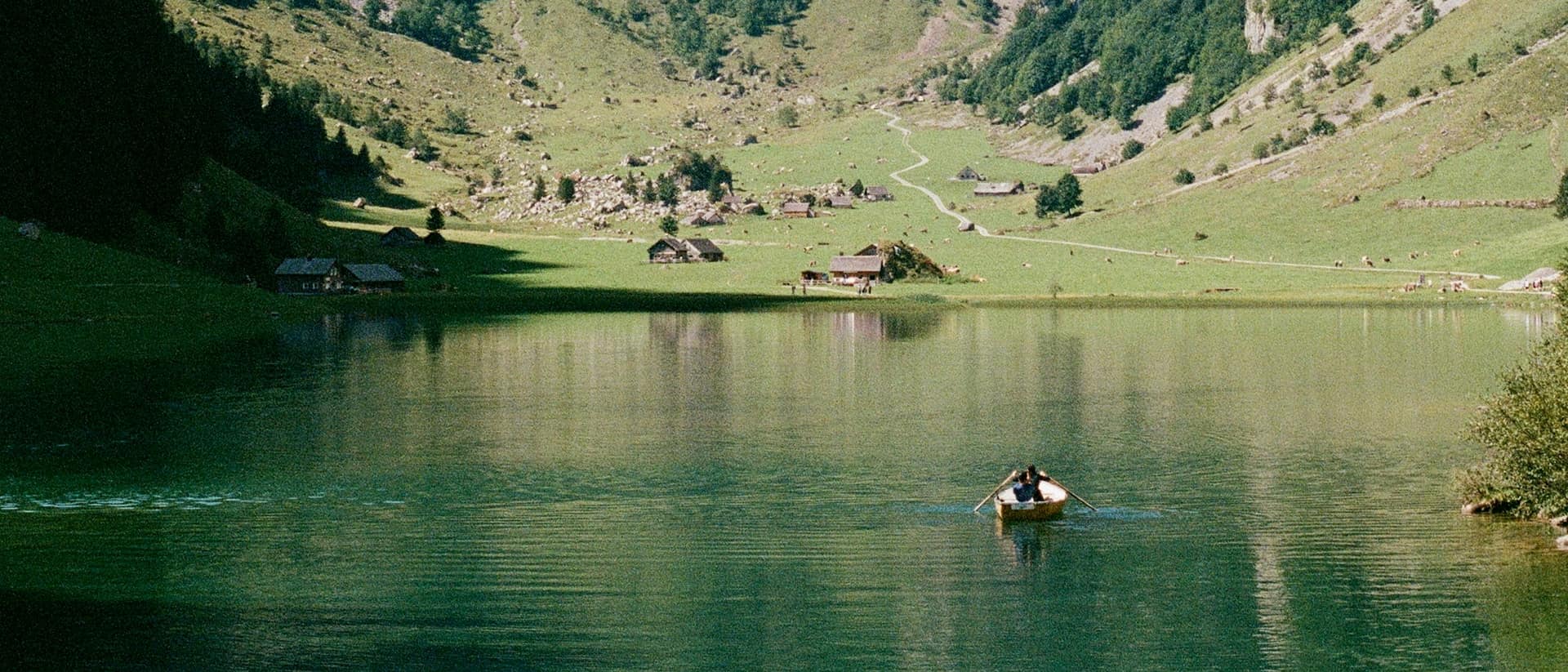 Ein Ruderboot auf dem Seealpsee im Alpstein, am Ufer Alphütten und grüne Weiden.
