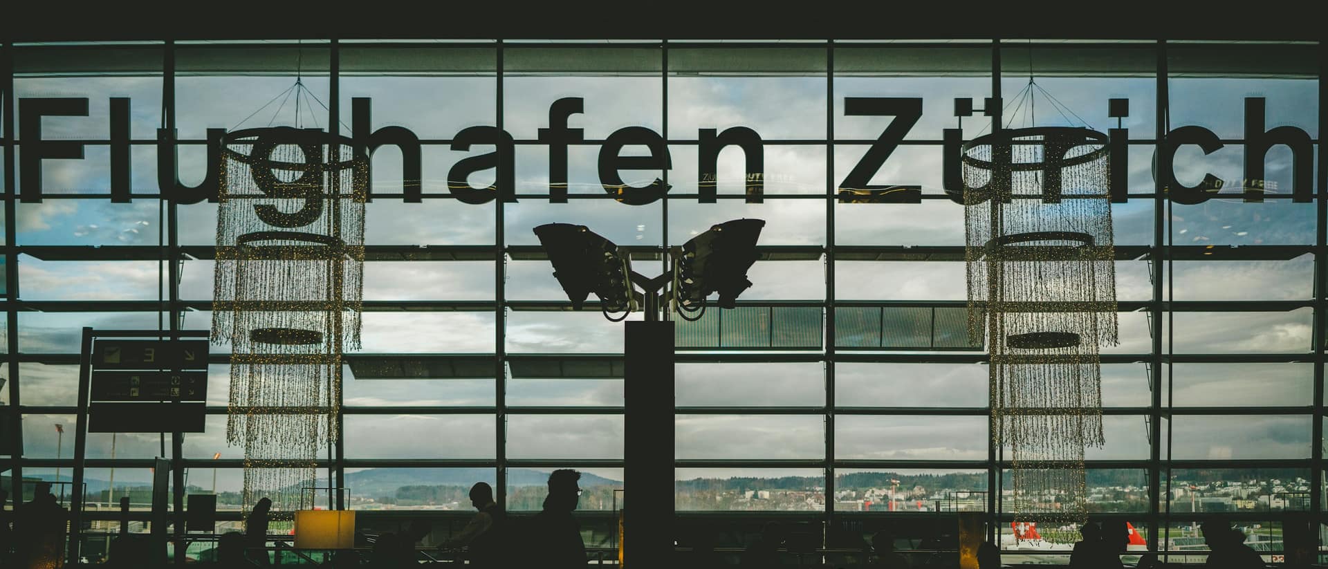 Pessoas à espera, em silhueta, diante da fachada de vidro do átrio de partidas com a inscrição Flughafen Zürich.