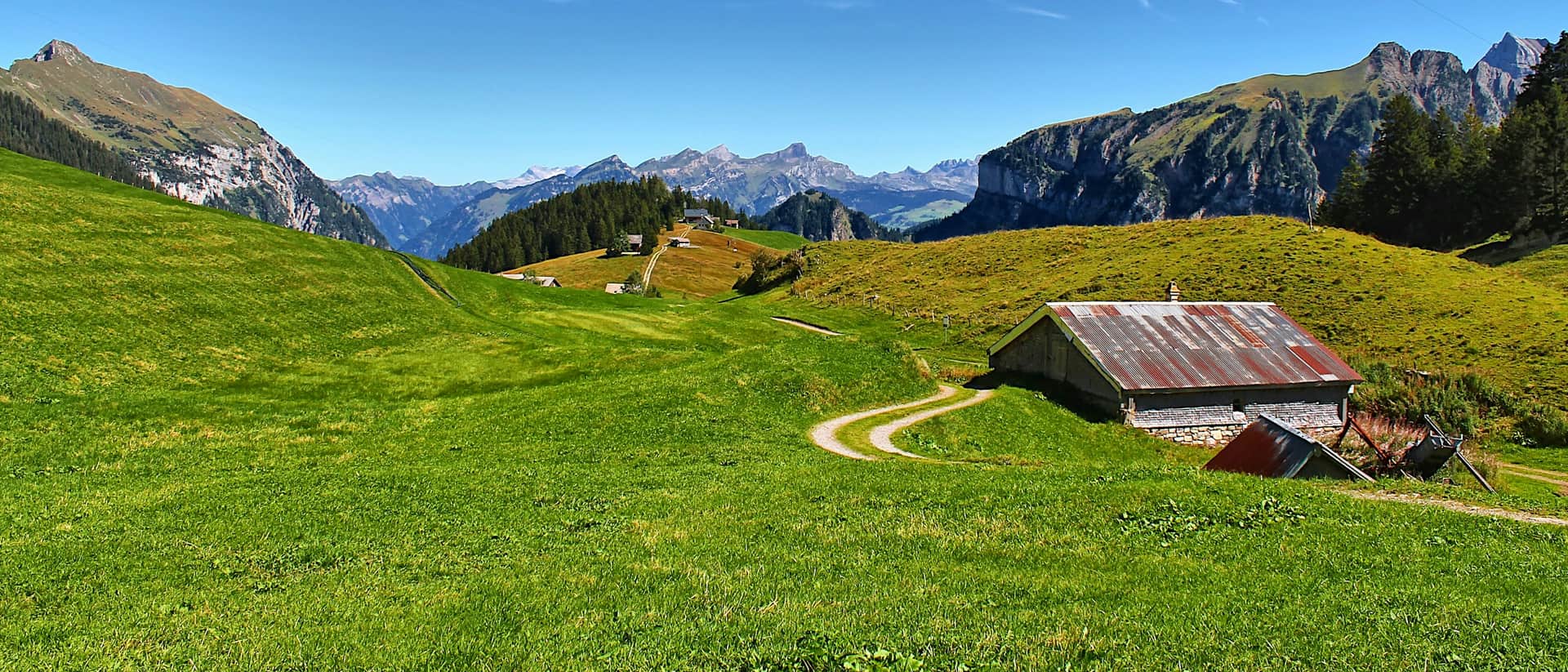 Ein alter Alpstall mit rostrotem Blechdach auf einer grünen Alpweide in Gitschenen, dahinter Bergketten unter blauem Himmel.