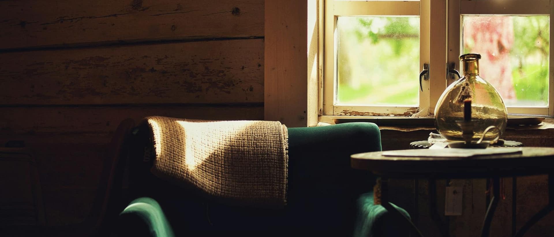 Ein Sessel mit Wolldecke steht in einer holzgetäfelten Stube am Fenster, durch das warmes Tageslicht fällt.