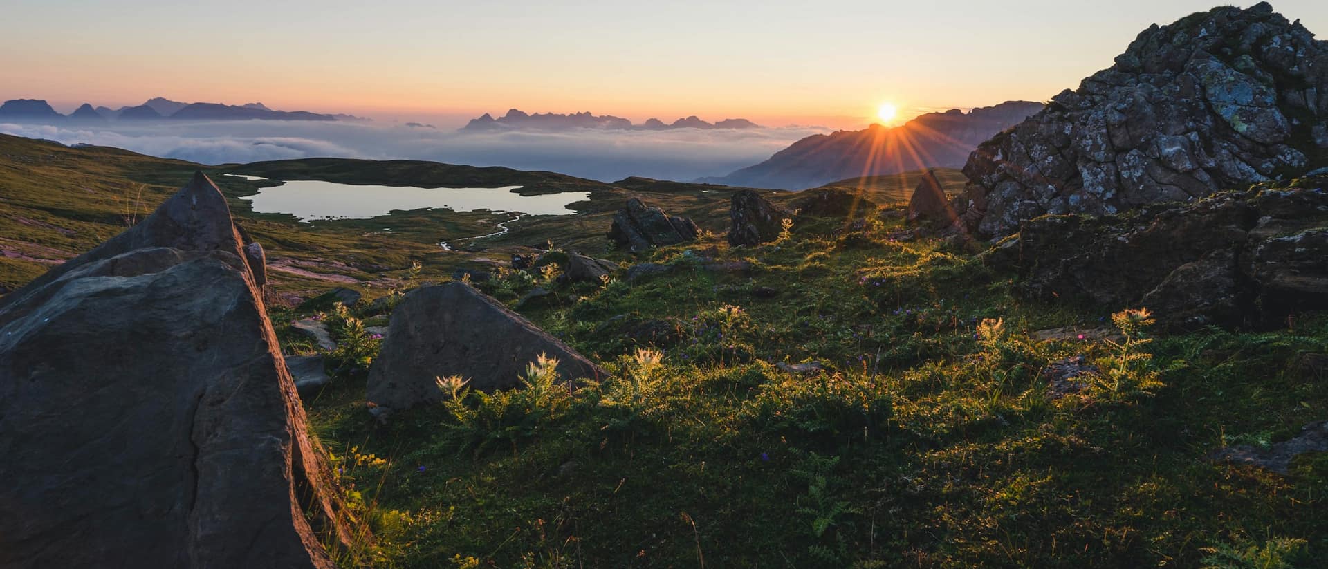 Sonnenaufgang über einer Alp bei Flums, im Vordergrund Felsen und Gras, in der Ferne ein Bergsee über dem Nebelmeer.